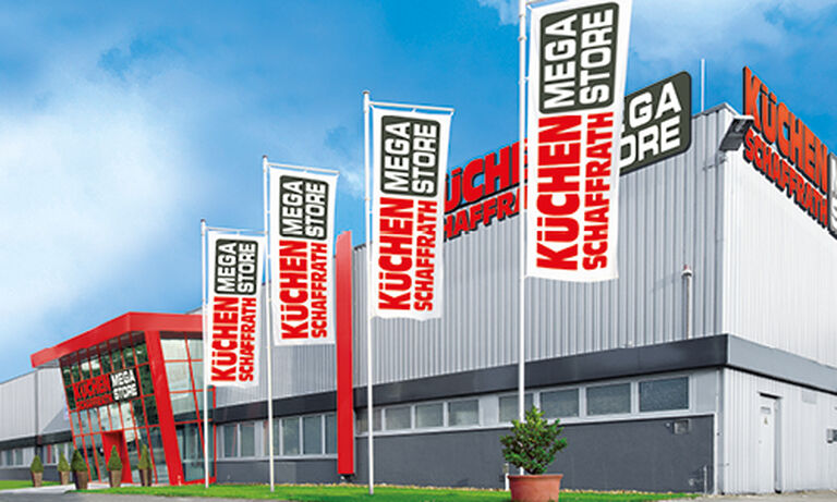 Das Bild zeigt den „Küchen Schaffrath Mega Store“ mit einer grauen Fassade und auffälligen roten Akzenten. Auf der linken Seite befindet sich ein Eingang mit großen roten Rahmen und Glasfronten, durch die der Innenbereich erkennbar ist. Entlang der Fassade stehen mehrere Fahnenmasten mit weißen Fahnen, auf denen in roten und schwarzen Buchstaben „KÜCHEN SCHAFFRATH MEGA STORE“ steht. Rechts oben auf dem Gebäude prangt ein großes Logo mit dem gleichen Schriftzug in roten und schwarzen Buchstaben.